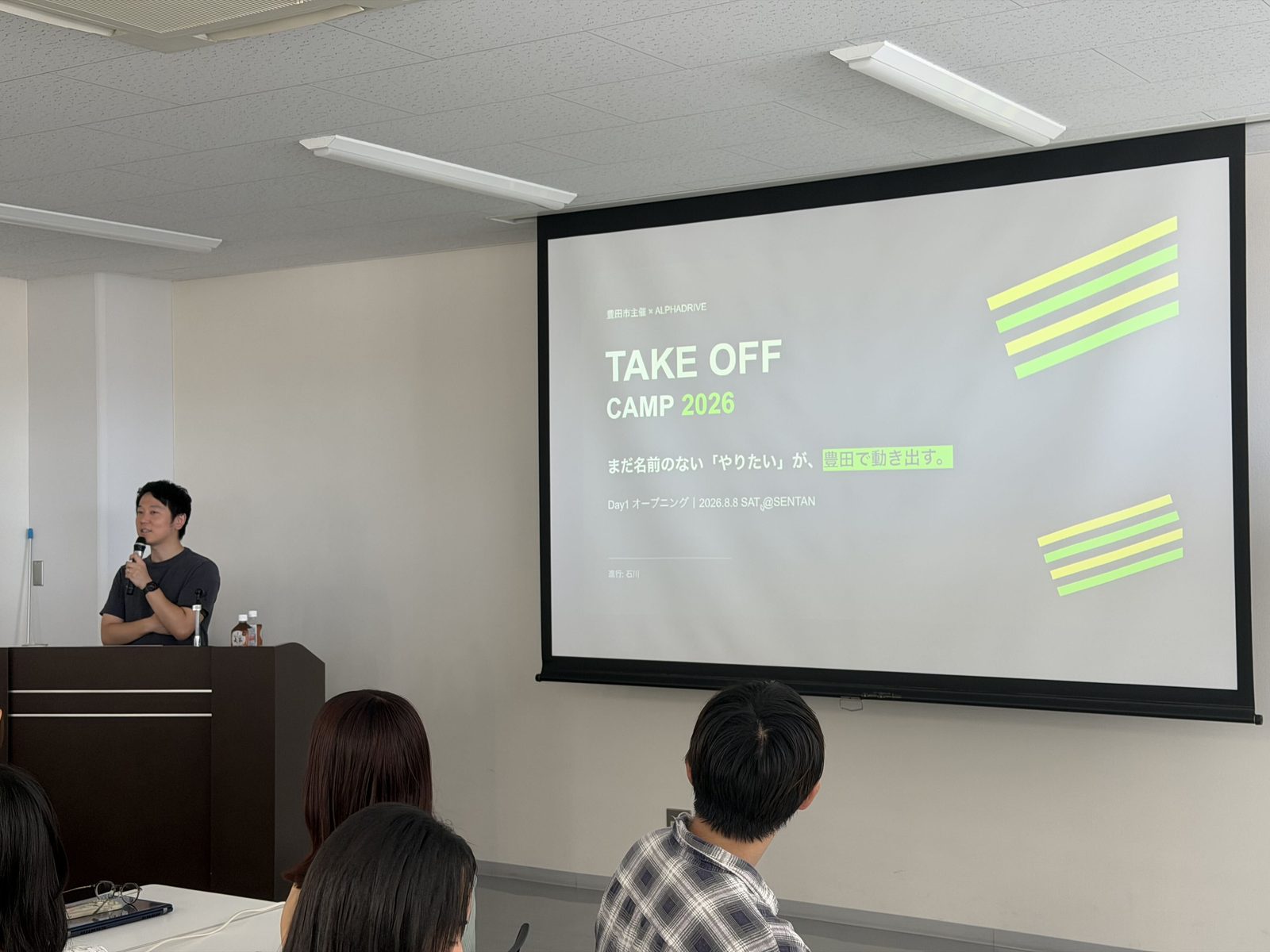 TAKEOFF Camp 2026のオープニングスライドを背に、マイクを持って話す様子