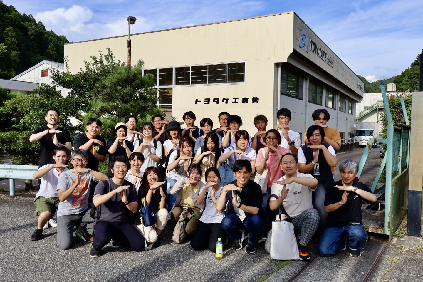 トヨタケ工業の社屋を背に、参加者と横田社長がTAKEOFFのTポーズをして並ぶ集合写真